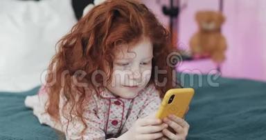 有趣的红头发小女孩与黄色智能手机躺在床上和使用智能手机。 沟通，播放，APP..